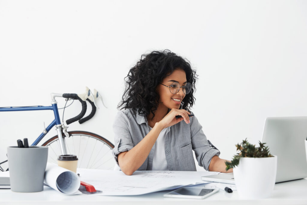 Happy pretty female engineer sitting over office interior having documents, gadgets and flowerpot on her desk checking her e-mails in front of open laptop. People, modern technologies, career concept